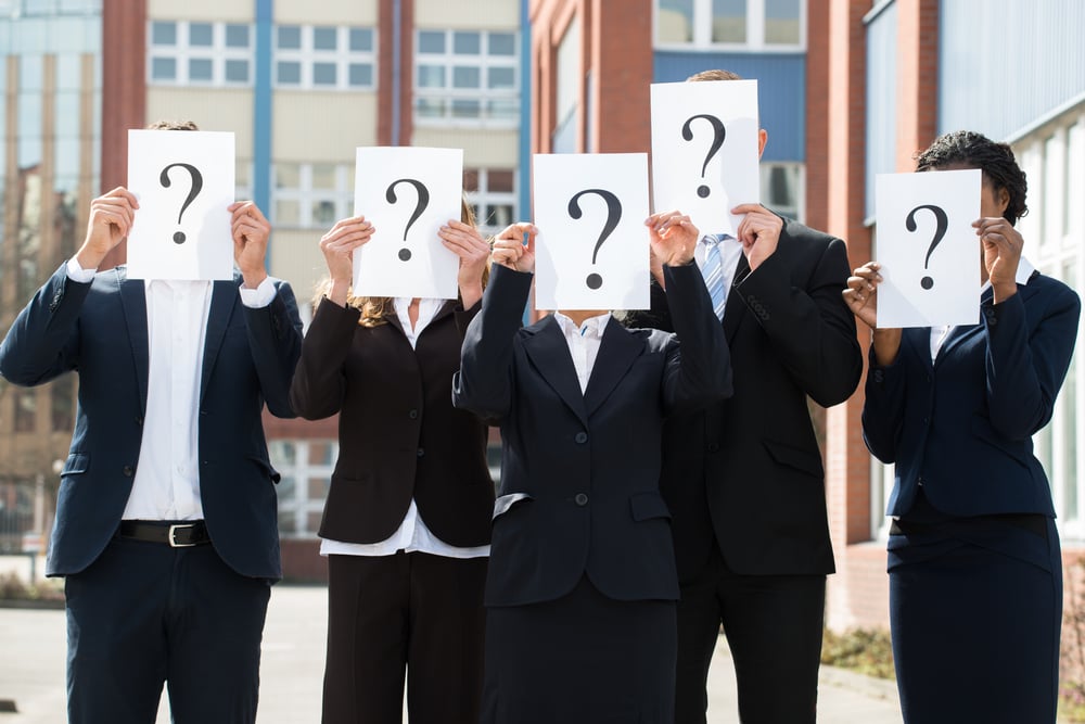 Five business people in formal office wear hold up sheet of paper with question mark in front of their faces